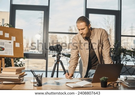 businessman in casual attire writing notes near digital camera and laptop on work desk in office