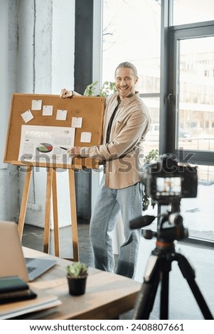 happy entrepreneur pointing at flip chart with analytics near digital camera in office, video blog