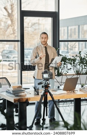 businessman in casual attire standing with document near digital camera and laptop, video marketing