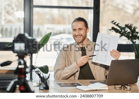 cheerful businessman showing blueprint in front of digital camera during video blog in modern office