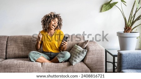 Excited happy young black woman holding smart phone device sitting on sofa at home - Happy satisfied female looking at mobile smartphone screen gesturing yes with clenched fist - Technology concept Royalty-Free Stock Photo #2408816721