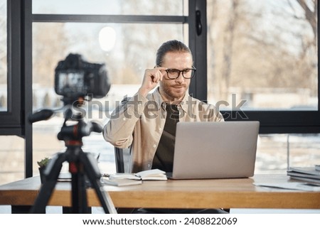 confident businessman in eyeglasses looking at laptop in front of digital camera in modern office