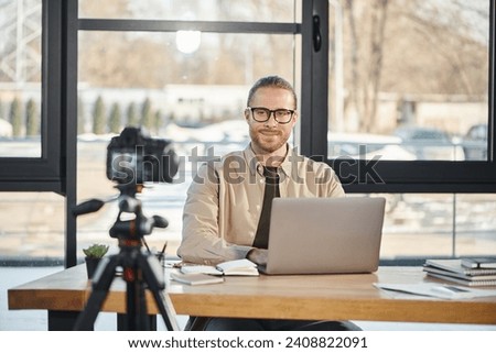 happy manager in eyeglasses sitting near laptop and smiling at digital camera during video blog