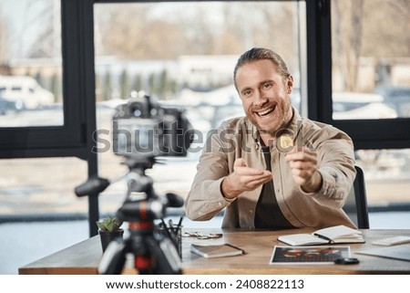 joyful successful entrepreneur showing bitcoin at digital camera and recording video blog in office