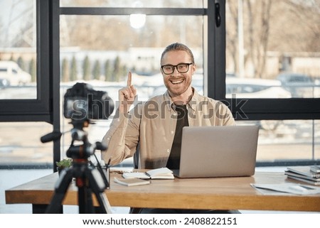happy businessman showing idea sign while sitting near laptop in front of digital camera, video blog