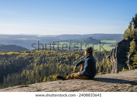 A middle-aged man enjoys the mountain landscape. Sits on the edge of the abyss. Elbe River Valley, Saxon Switzerland National Park, Germany. Active lifestyle Royalty-Free Stock Photo #2408833641