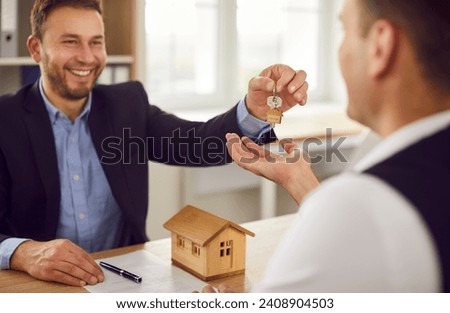 Happy, friendly man realtor or real estate agent sitting at his office table with a contract and a house model, smiling and handing keys to the buyer. Real estate, property purchase concept  Royalty-Free Stock Photo #2408904503
