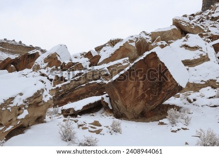 Snowy picture of red rock boulder that has ancient writings on it of animals. 