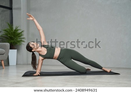 Graceful fit female in sportswear doing side plank exercise, while training at home. Front view of beautiful woman in sports bra standing side forearm plank during yoga session. Concept of yoga. Royalty-Free Stock Photo #2408949631