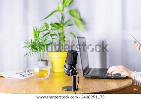 Woman preparing to make podcast in her home studio with laptop, microphone, cup of herbal tea and bright potted flowers on the table.