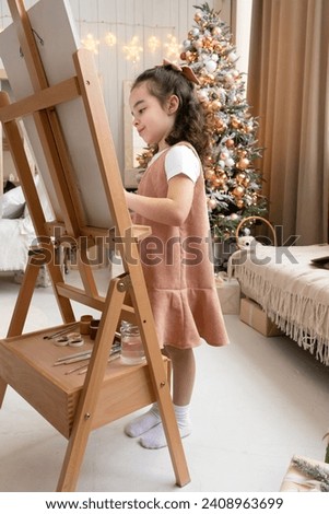 Smiling cute girl paints on easel with palette and paintbrush. Art school. Christmas tree background. Merry Christmas and Happy New Year Eve. Vertical photo.