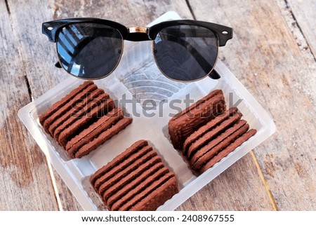 Stack of creamy and tasty chocolate cookie biscuits in a plastic container with black sunglasses over wooden background