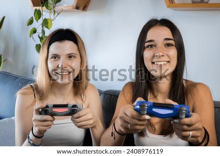 two friends laughing and playing video games. in the living room sitting on their couch. enjoying free time together. both wearing a white t-shirt and black shorts.
