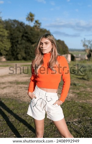 Fashion Photography: Woman in the middle of a rural field, standing in an area with green grass and an earthy path. Wearing an orange sweater and beige long shorts. Sunny spring summer day. Lifestyle