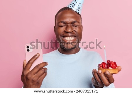 Cheerful dark skinned bearded man wears cone hat smiles broadly holds delicious strawberry cupcake and smartphone receives congratulations on birthday isolated over pink background. Festive occasion