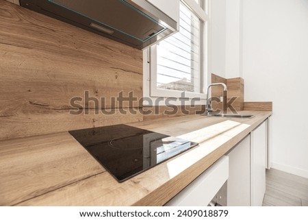 An open kitchen with white cabinets and wood countertops with matching backsplash with small black cooktop and white aluminum window Royalty-Free Stock Photo #2409018879