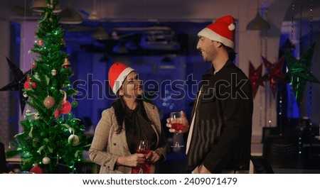 Two Indian colleague in santa hat standing at indoor office hold glass drinking juice toast together enjoying night new year 2024 party. Happy smiling beautiful female young male make funny gossips