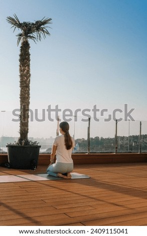 a dark-haired girl meditating in a girlish way and dressed in sports clothes sits on a fitness mat on a wooden street terrace against a background of a blue sky, a palm tree is placed nearby