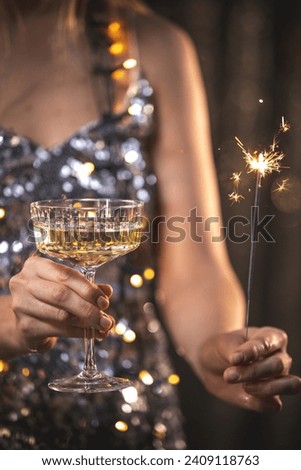 The girls pour champagne into round glasses from a bottle. Women in a sparkling dress. Festive decoration, Christmas garland. Full cocktail creamers - le coupe. The female's hand holds a wine glass