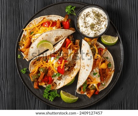 baked tex-mex chicken fajitas with mixed sweet pepper, onion, sour cream, shredded cheese and white corn tortillas on black plate on black wooden table, horizontal view, flat lay, close-up
