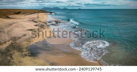A panoramic aerial view of Sandy Beach in São Vicente, Cape Verde, captures the tranquil waves washing over the golden sands, contrasted by the dramatic outline of distant islands under a dynamic sky