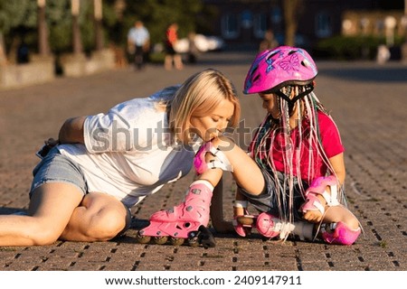 Mother kisses her little daughter's injured knee. Mom is teaching her little daughter to ride roller skates in summer park