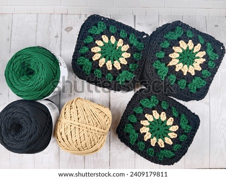 A picture of crochet granny squares in green, black, and cream colors on some white table in a house. They can be joined to be a tablecloth, table runner, even some cardigan.