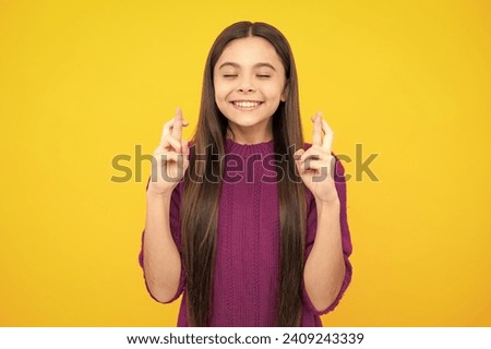 Teenager child holding fingers crossed for good luck. Teen girl prays and hopes dreams come true, isolated on yellow background.