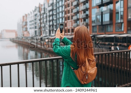 Back view of young European tourist makes photo or video on smartphone at city of Gdansk, Poland. Hipster girl with backpack hold on smart phone gadget outdoors