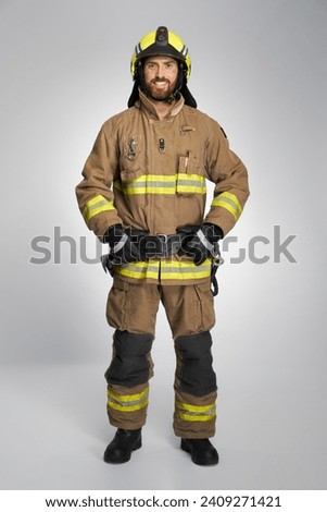 Smiling bearded fireman in protective uniform posing in studio. Front view of happy caucasian male rescuer keeping hands on belt, while looking at camera, on gray background. Emergency job concept.