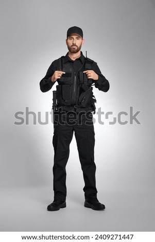 Serious male policeman in full gear standing, posing indoors. Front view of caucasian cop keeping hands on body armor, while looking camera, on gray background. Concept of work, equipment, safety.
