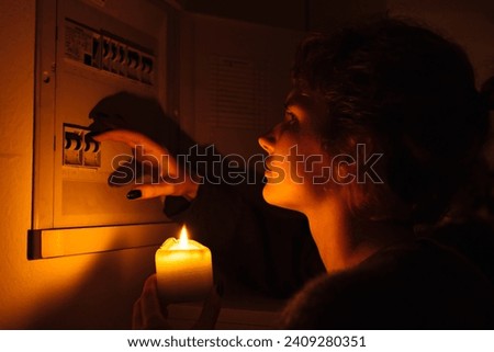 young woman with candles in complete darkness examines fuse box or electrical distribution board at home during power outage. Blackout, no electricity Royalty-Free Stock Photo #2409280351
