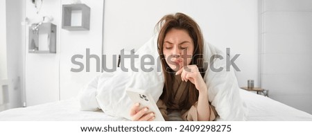 Image of young sad woman with smartphone, lying in bed wrapped in blanket.