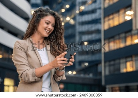 Happy smiling confident businesswoman standing in front of the modern workplace in big city using smartphone for texting, cellphone apps for work outside. Royalty-Free Stock Photo #2409300359
