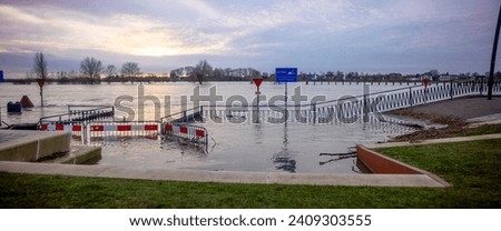 Barrier fence under water at flooded countenance boulevard of Zutphen with high water level of river IJssel reaching walking path. Panoramic view. Royalty-Free Stock Photo #2409303555
