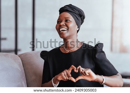 Joyful woman in black turban and dress smiling and creating a heart shape with her hands