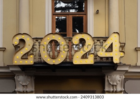 Happy New Year 2024. Volumetric letters with light bulbs. Text. Golden numbers 2,0,4 on balcony of old yellow house at city festive Christmas market of Tbilisi. Winter holiday concept. Wooden door
