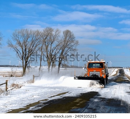 Orange snowplow a on beautiful sunny day, throwing up snow as it plows a country road after a snow storm Royalty-Free Stock Photo #2409335215