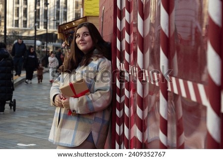 Street photo. Lonely young beautiful white woman with craft gift box at Christmas market leaning on wall with red and white stripes. Joyful happy childless brunette is thinking about New Year 2025