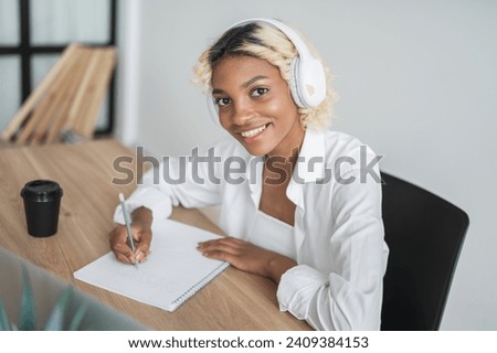 Happy college student girl looking at the camera wears headphones and taking notes. Prepapre for exam,lecture,report etc Royalty-Free Stock Photo #2409384153