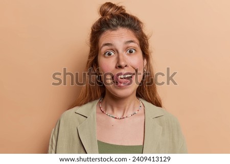 Photo of funny European woman with long hair sticks out tongue and foolishes around makes funny expression focused at camera wears shirt isolated over brown background notices something tasty
