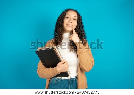 Happy Student arab girl with curly hair with toothy smile, keeps index fingers near mouth, fingers pointing and forcing cheerful smile