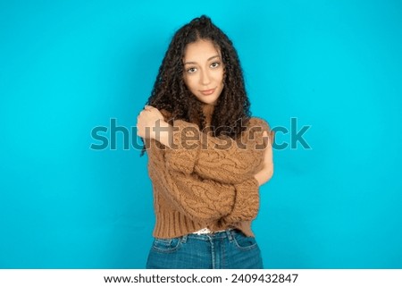 Charming pleased Beautiful arab teen girl with curly hair wearing knitted sweater embraces own body, pleasantly feels comfortable poses. Tenderness and self esteem concept