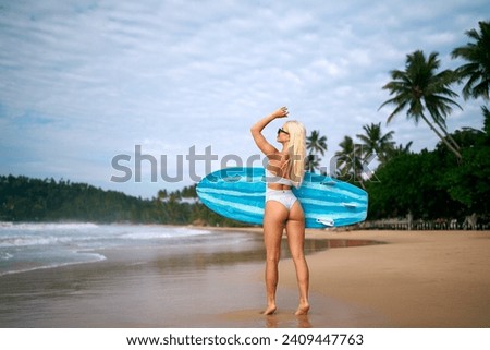Blonde woman in bikini holding surfboard on tropical beach, preparing for surfing session. Female surfer enjoying summer vacation by the sea, getting ready to catch waves under sunny sky.