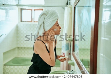 Woman in a vintage bathroom applies sunscreen to her face for skin protection. Towel-wrapped hair, retro tiles, skincare routine, mirror reflection capture the essence of daily beauty regimen. Royalty-Free Stock Photo #2409447805