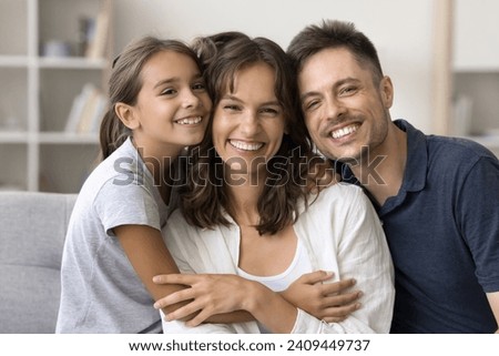Cute loving daughter kid and happy father hugging beloved mom with gratitude, tenderness, affection. Parents and child sitting on home couch with toothy smiles, posing for home family portrait