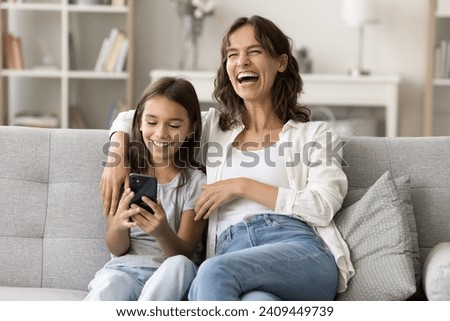 Cheerful excited mom and daughter child enjoying Internet technology on smartphone, having fun, laughing out loud, watching funny content, taking family selfie picture on mobile phone
