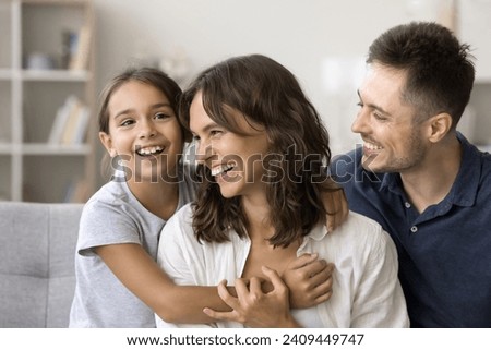 Carefree cute little girl kid hugging positive mom and dad on couch, smiling, laughing. Happy couple of parents playing with child on sofa, enjoying care, family leisure time at home