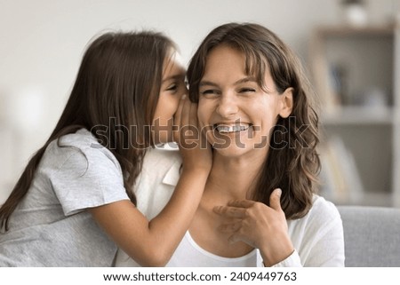 Positive cute girl telling secret to happy mom, whispering at ear. Cheerful mother enjoying family communication with child, motherhood, family time listening to kid laughing Royalty-Free Stock Photo #2409449763