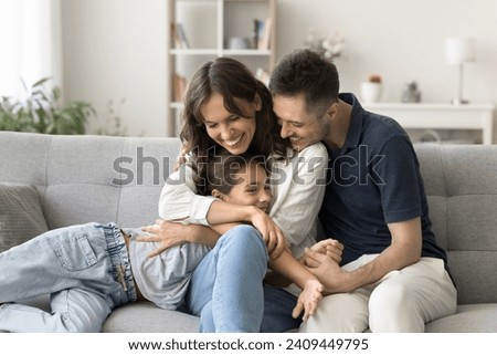 Happy young parents cuddling sweet adorable kid girl on comfortable home couch, hugging child with love, adoration, laughing, spending family leisure with daughter child together